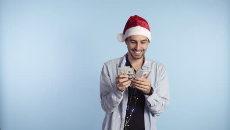 Young-happy-man-received-paper-money,-dollar-banknotes.-Counting-money-while-standing-against-the-blue-wall-in-santa-hat-and-garland-on-neck.-Get-money-cash,-receive-winner-payment,-gift-concept.-Man's-hand-hold-and-counting-cash-money-and-showing-as-fan