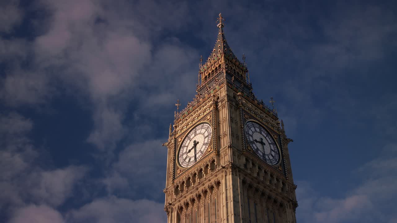 Majestic upward angle captures the ornate details of Big Ben’s clock faces and stonework, illuminated by sunlight and framed by swirling clouds above the iconic tower in London