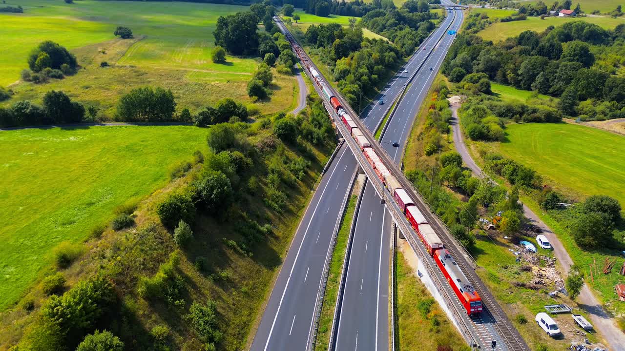 Luftaufnahme eines roten Güterzugs, der in einer malerischen Landschaft eine Brücke über eine viel befahrene Autobahn überquert