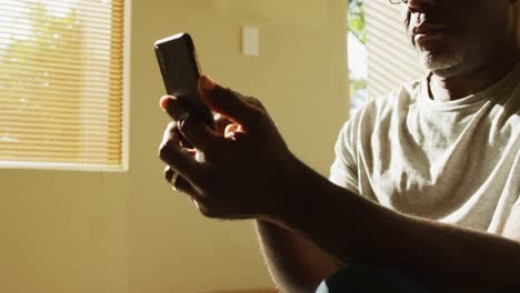 Un-Hombre-Mayor-Afroamericano-Pensativo-Sentado-En-Casa-Bajo-El-Sol-Usando-Un-Teléfono-Inteligente