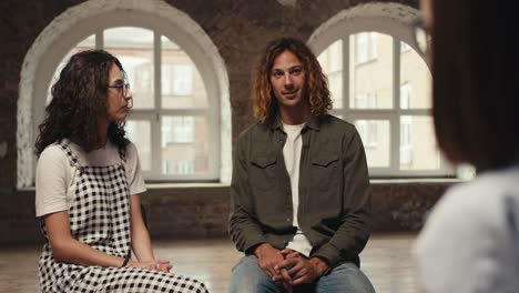 A-brunette-girl-with-curly-hair-with-glasses-in-a-checkered-jumpsuit-and-a-brunette-guy-in-a-green-jacket-communicate-with-a-professional-psychologist-with-a-bob-haircut-and-glasses-during-therapy