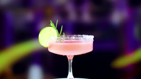 a pink margarita cocktail garnished with lime and rosemary, set against a blurred, colorful background. bright lighting enhances the drink's appeal