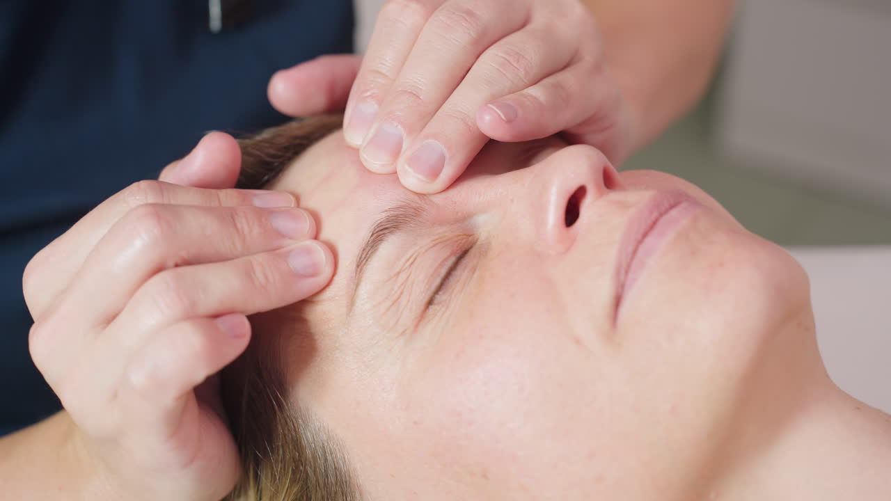 Facialist massaging face of client, patient tenses slightly then relaxes under gentle fingertip pressure, close up focus on tension release around brow region, spa ambient, spa lighting