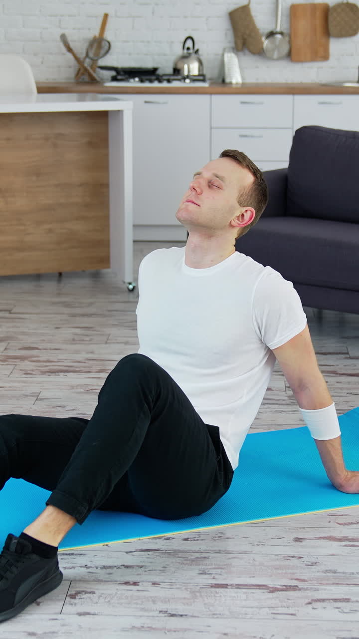 Man relaxes on a mat indoors. Young man sitting on a mat after hard workout at home. Fitness during quarantine.. Vertical video