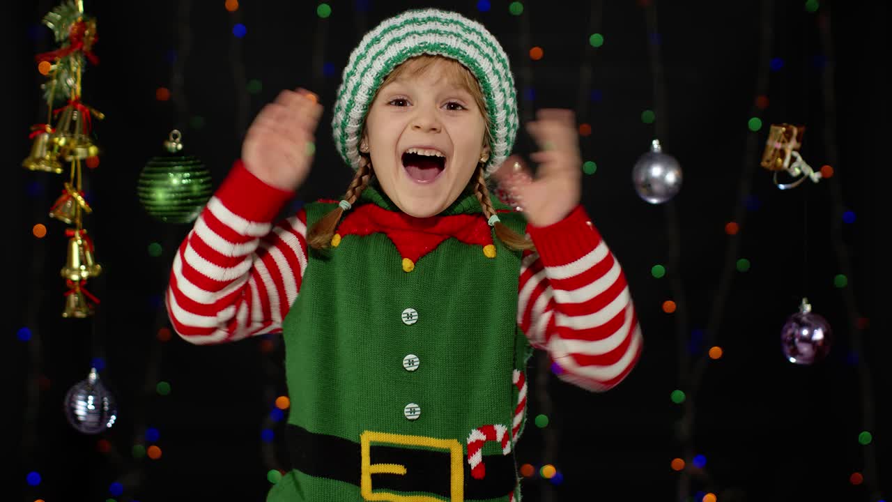 Holčička v kostýmu vánočního skřítka, pomocníka Santa Clause. Dítě dělá vítězné gesto, mává rukama
