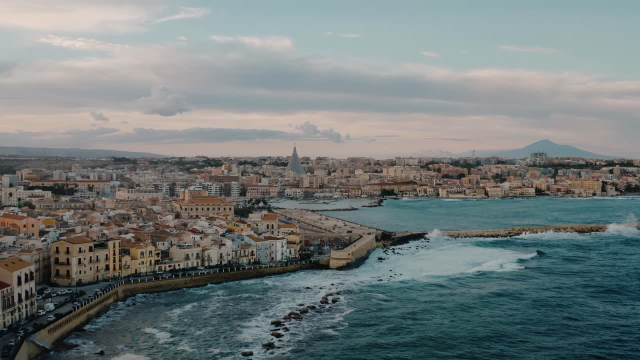 Aerial view of Ortigia island and Mount Etna in a distance. Wide drone shot over historic buildings by Mediterranean sea with waves crashing on the shore in the evening. UNESCO. Sicily. Italy.