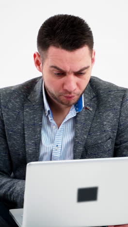 grieved businessman looking into the laptop. pensive male freelancer working on a modern gadget while sitting alone on white background. vertical video