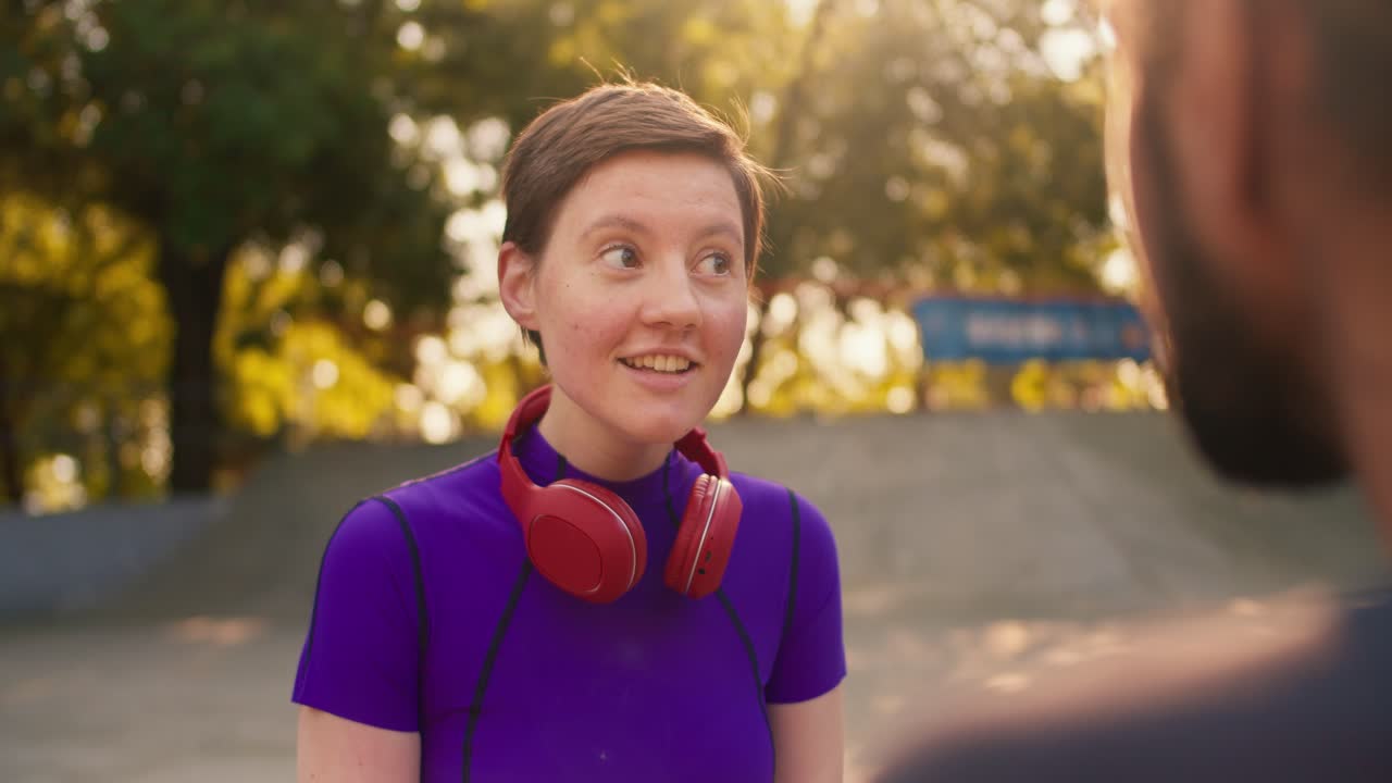vista en primera persona: un tipo moreno con una camiseta gris se comunica con una chica con un corte de pelo corto en una camiseta púrpura y auriculares rojos en un parque de skate en verano