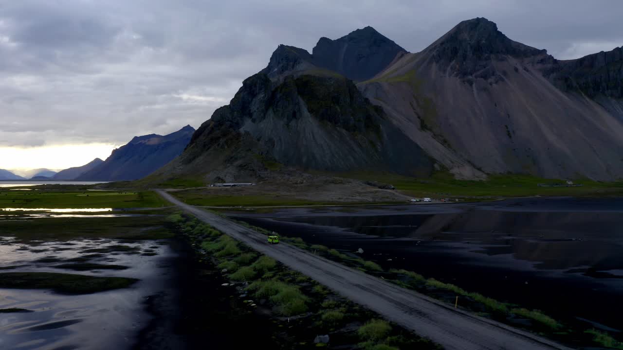 Drone shot of a car driving through dramatic landscape of Vestrahorn mountains and beach under an overcast sky