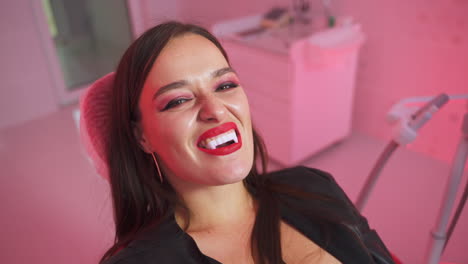 dark-haired woman with vampire teeth sits at dentist's chair. dentist cabinet lit with red light at backdrop.
