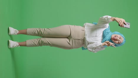 full body of asian muslim woman smiling and using smartphone taking picture while standing in the green screen background studio