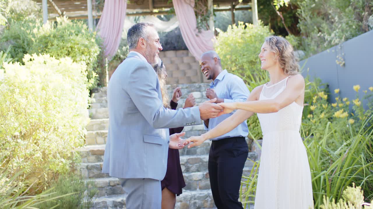 Una coppia mista in smoking e abito da sposa si abbraccia e ondeggia sui gradini di pietra mentre gli invitati applaudono, irradiando gioia.