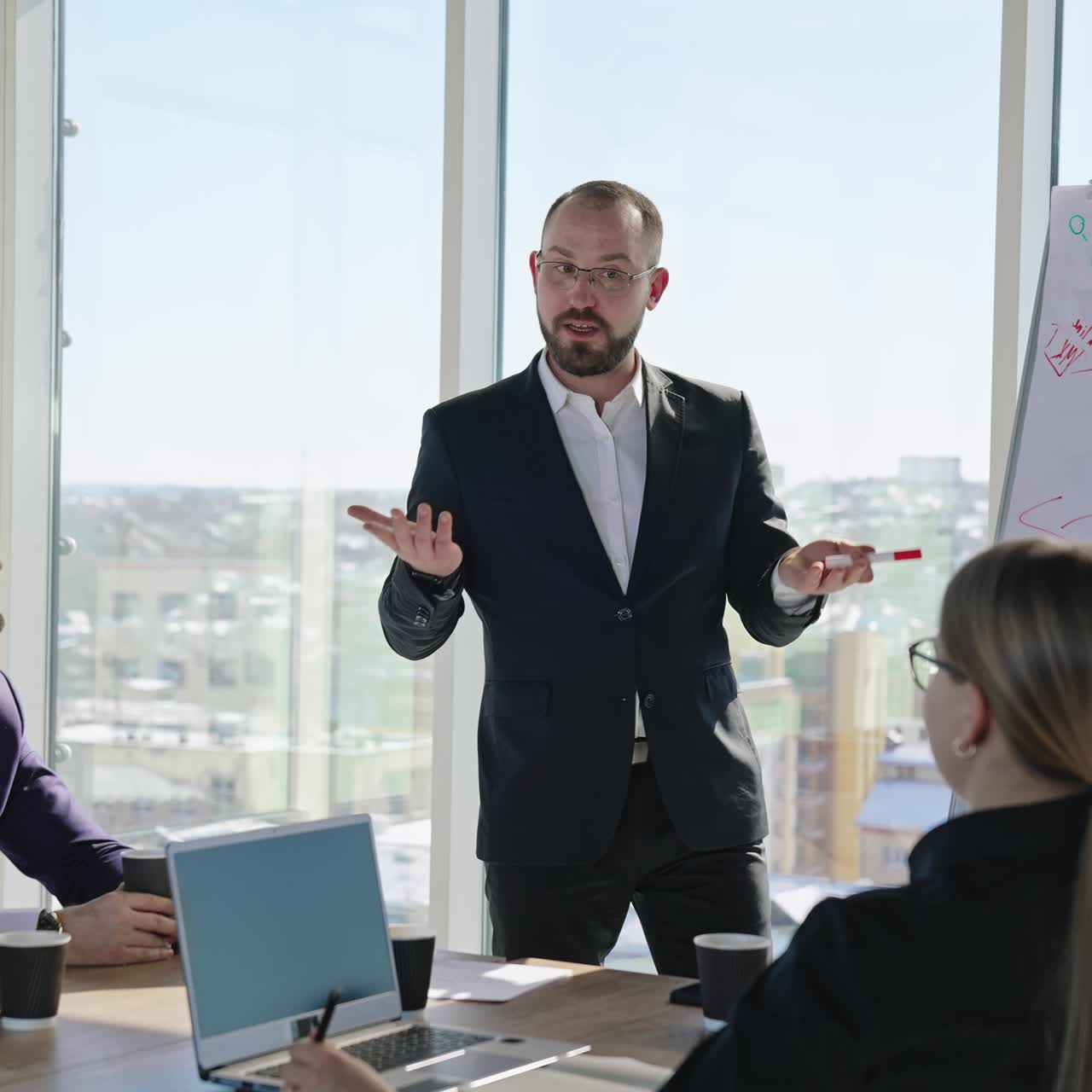 Office workers sit at the table with laptops and look at the speaker. The presenter gives a talk emotionally in front of the colleagues. Business presentation in the light office