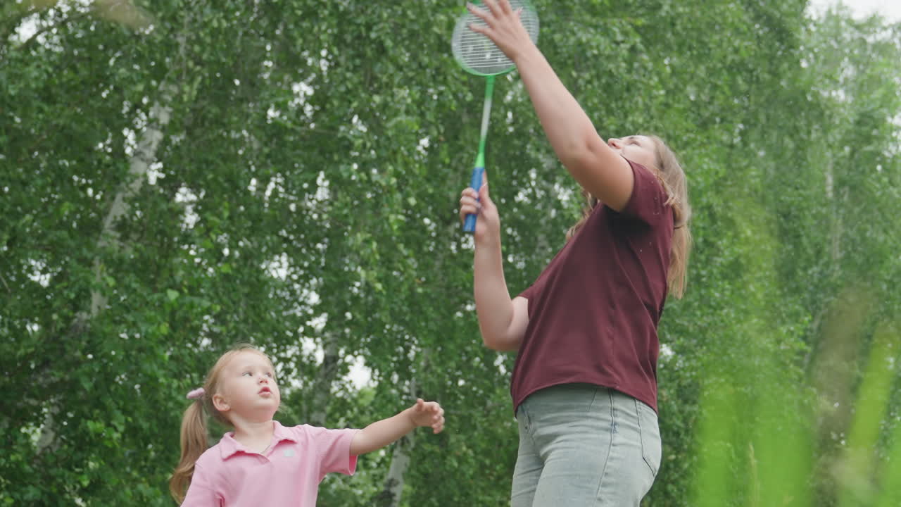 caucasian mother teaching daughter badminton outdoors mother coaches child with racket and shuttlecock under green birch trees casual clothing sunny afternoon gentle guidance playful laughter