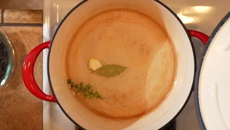 heating and seasoning water with a sprig of rosemary, onion and a bay leaf - lifting the lid off the pot in overhead view