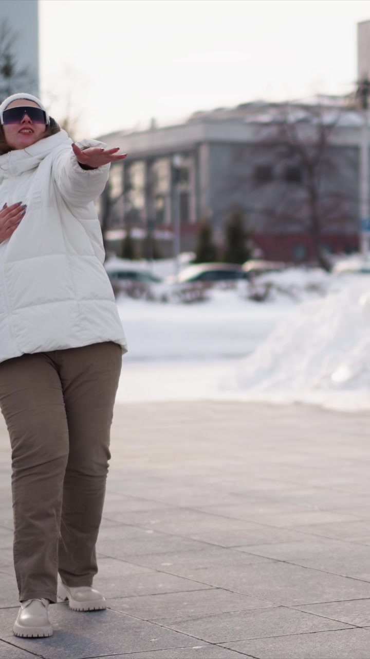 Eine Frau tanzt im Freien in einer verschneiten Stadtumgebung, trägt eine weiße Steppjacke, beige Hose und Kopfhörer. Die Frau bewegt sich energiegeladen über den gepflasterten Boden inmitten schneebedeckter Umgebung
