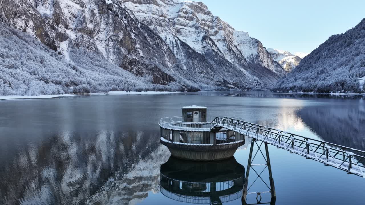 Klöntalersee, Glarus, Switzerland, featuring a modern pumphouse structure on the dark, reflective lake. Snow-covered mountains frame this blend of nature and utility
