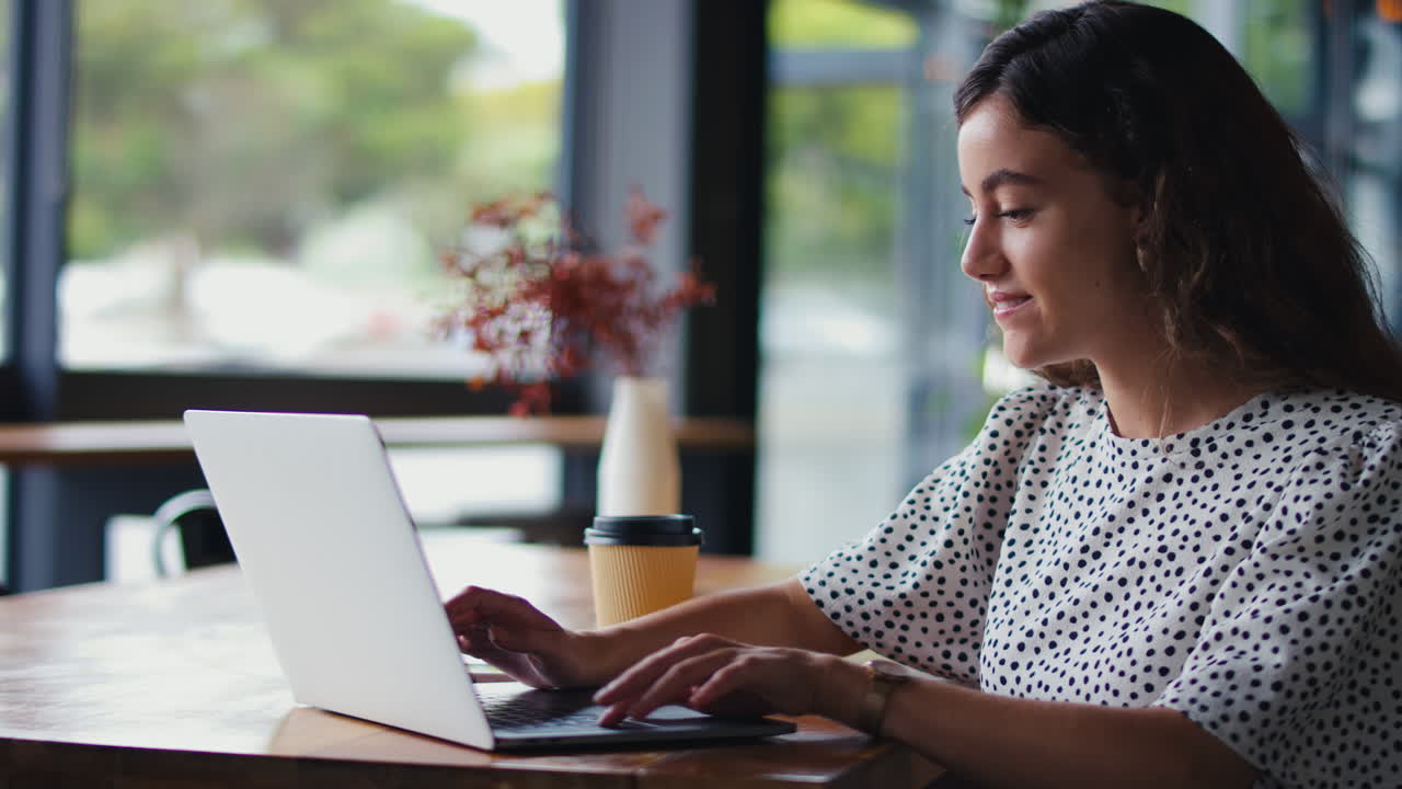 Young Businesswoman Sitting With Laptop In Coffee Shop Taking Call On Mobile Phone Using Microphone