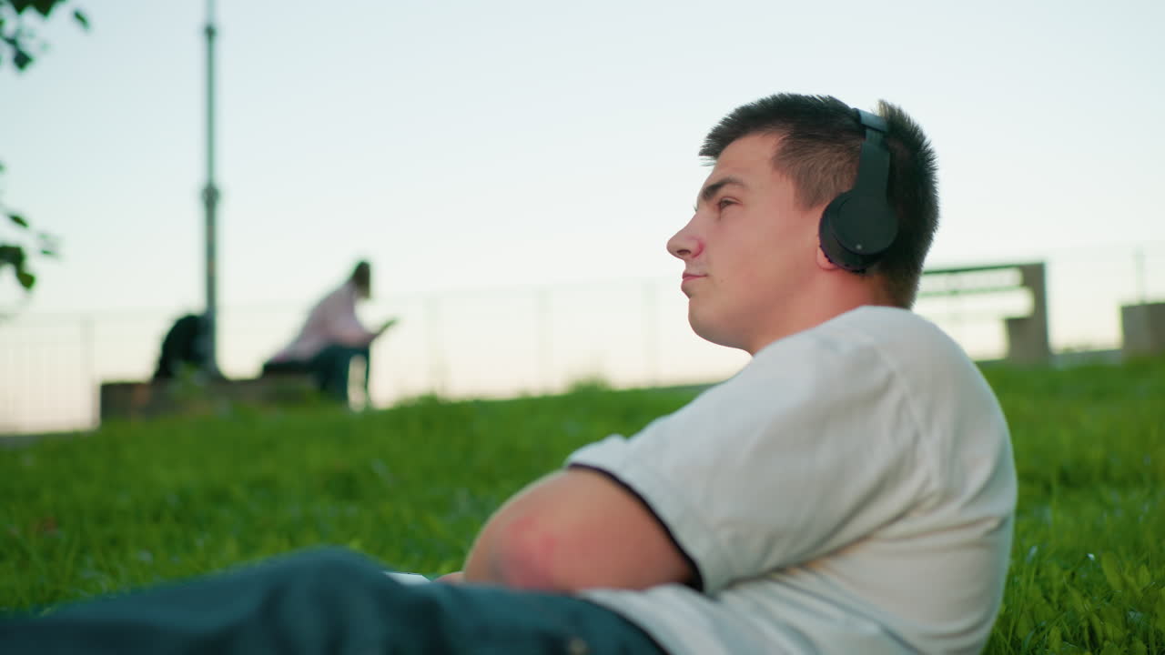 남자는 머리를 지하는 손으로 잔디에 으로 누워서 헤드폰을 착용하고 음악을 즐기고, 누군가는 멀리 배경에 앉아서 흐릿하게 보입니다.