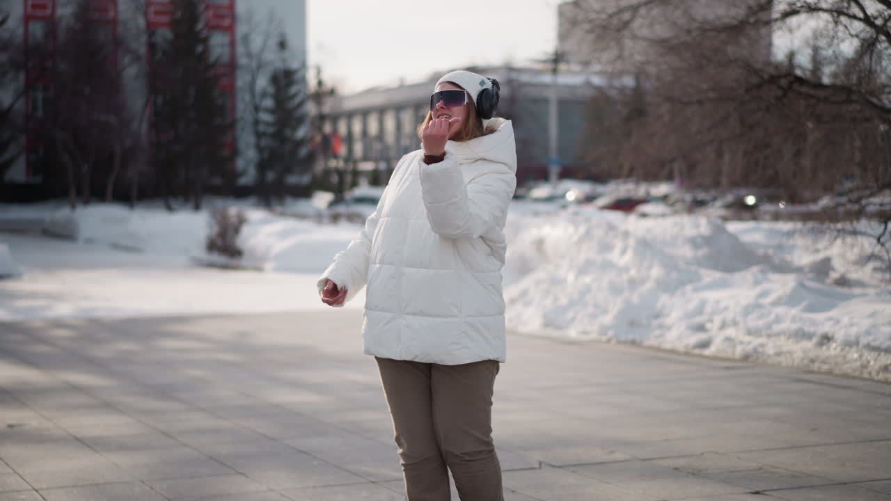 School girl in white winter coat, beanie and headphones dancing, talking and demonstrating moves on paved recreation park walkway with snow-covered ground, passing pedestrians and trees