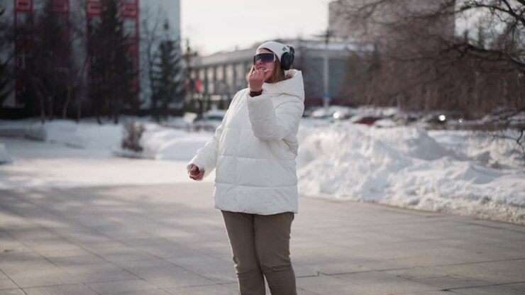 School girl in white winter coat, beanie and headphones dancing, talking and demonstrating moves on paved recreation park walkway with snow-covered ground, passing pedestrians and trees