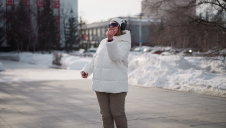 school girl in white winter coat, beanie and headphones dancing, talking and demonstrating moves on paved recreation park walkway with snow-covered ground, passing pedestrians and trees