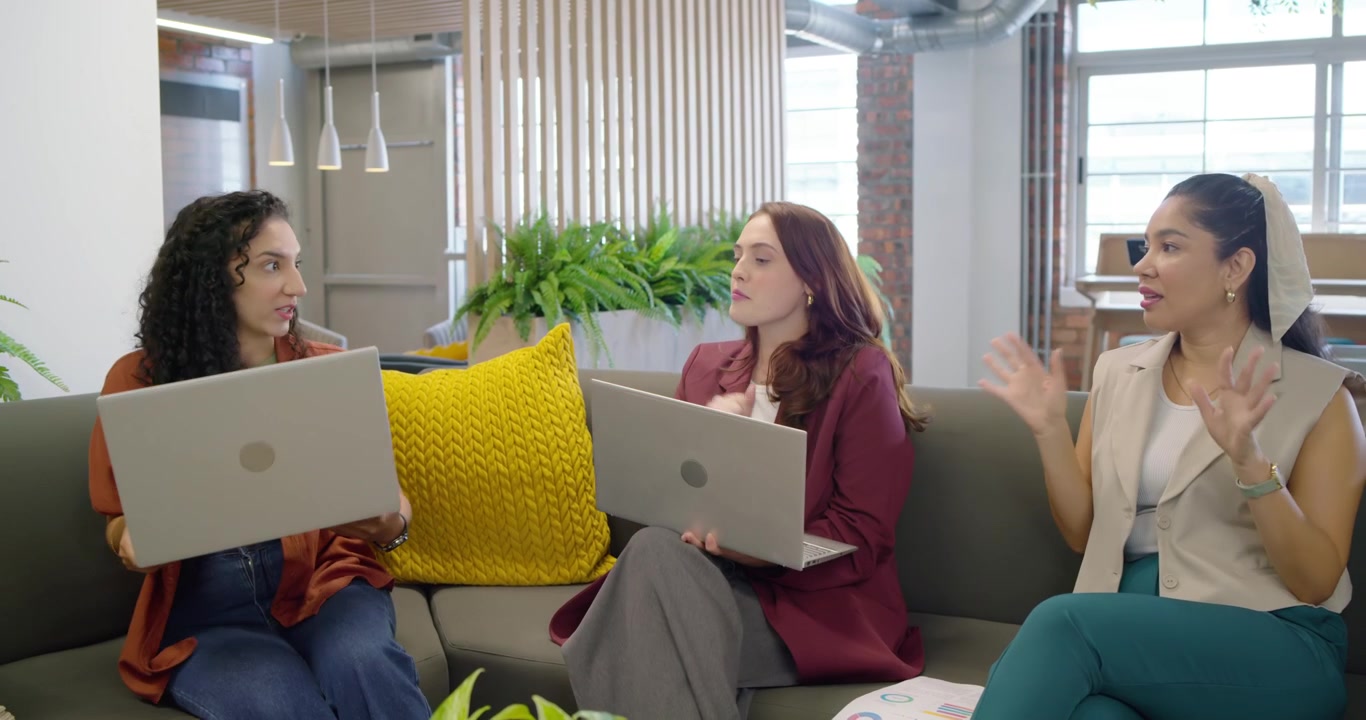 Female colleagues reviewing laptop and chart on sectional sofa in office lounge, passerby joining