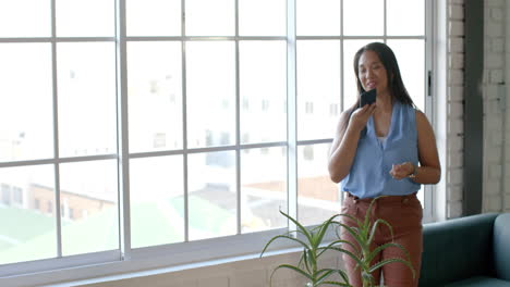 Biracial-woman-speaks-on-the-phone-by-a-large-window,-with-copy-space