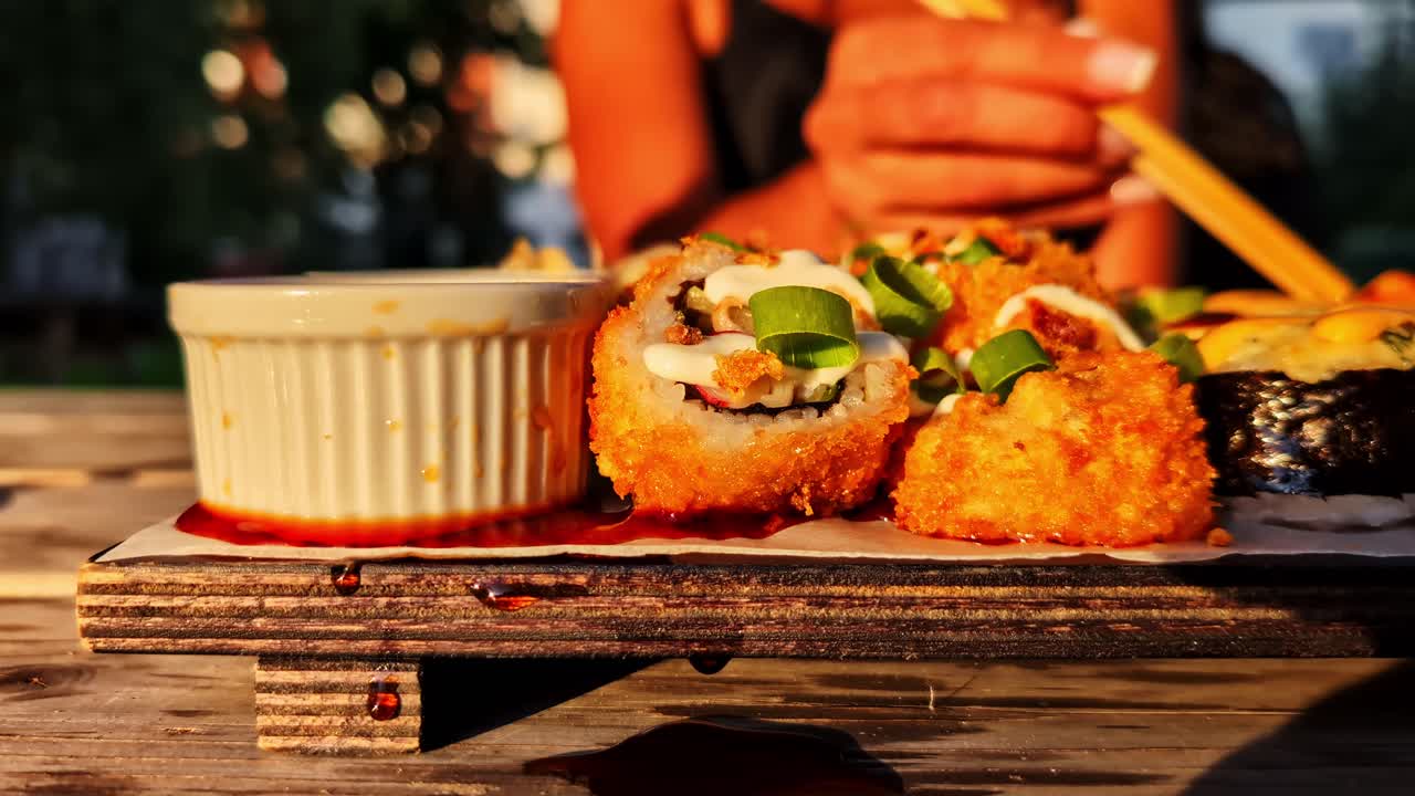 Static closeup of woman eating sushi, chopsticks lifting roll in golden outdoor lighting, dipping into soy sauce