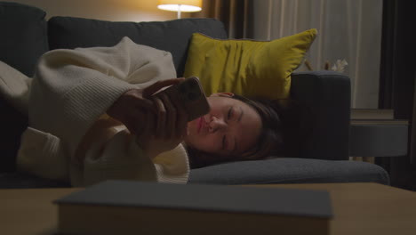 woman spending evening at home lying on sofa with mobile phone scrolling through internet or social media 7