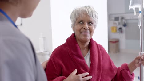 Happy-diverse-female-nurse-talking-to-senior-patient-with-drip-in-slow-motion,-unaltered