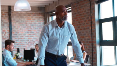 Portrait-Of-Serious-Mature-Businessman-Standing-In-Busy-Office-With-Colleagues-Working-In-Background