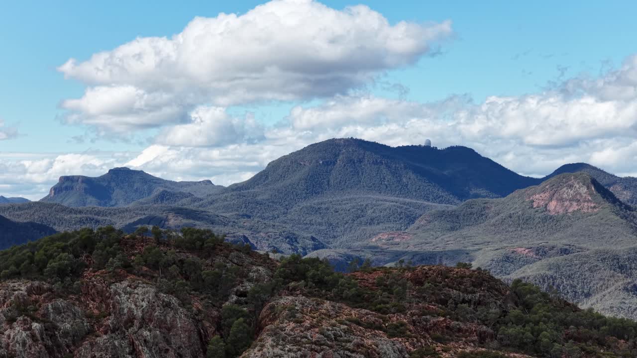 Slow camera pan reveals rugged Warrumbungle mountain range with scattered clouds, rocky outcrops, and forested hills in bright natural daylight