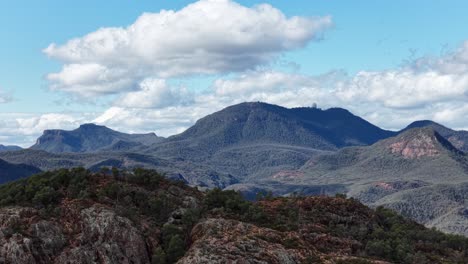 slow camera pan reveals rugged warrumbungle mountain range with scattered clouds, rocky outcrops, and forested hills in bright natural daylight