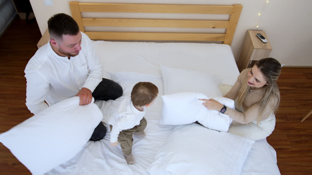 Loving parents sitting on the bed hit their little baby with pillows playfully. Kid with a pacifier in mouths laughs and likes a game. Top view.