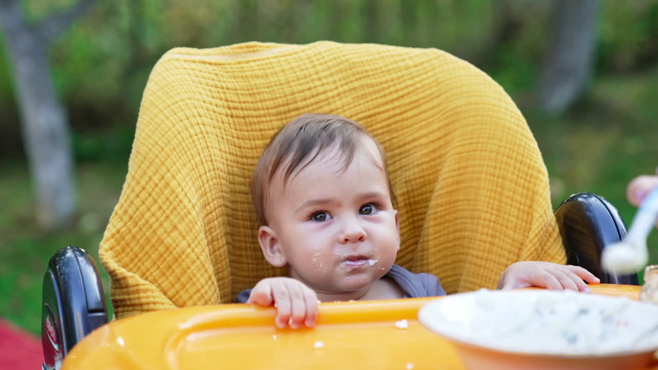 Cute little baby boy in a yellow chair being fed. Mom fills the spoon in plate and gives it to her child. Blurred backdrop.