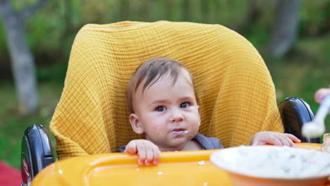 cute little baby boy in a yellow chair being fed. mom fills the spoon in plate and gives it to her child. blurred backdrop.