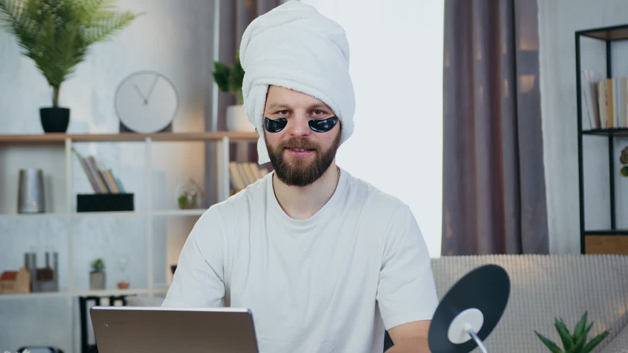 Attractive smiling modern young bearder looking into camera with collagen refreshing eye-patches and towel around his head,beauty concept