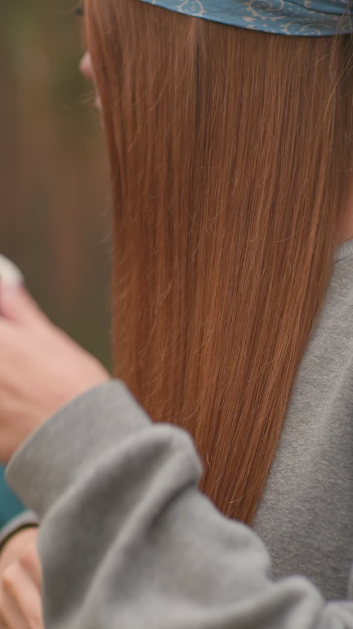 Close-up of woman brushing her hair, focusing on her long reddish-brown hair and gentle self-care moment, while another woman in the background, slightly blurred, opens her thermos