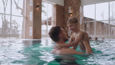 Gente-Feliz-En-El-Moderno-Centro-De-Bienestar,-Padre-E-Hijo-Pequeño-Nadan-En-La-Piscina-El-Fin-De-Semana-Familiar-En-El-Complejo-De-Atención-Médica