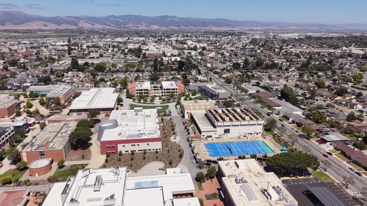 Drone video shows Hartnell College campus, modern buildings, athletic facilities, and Salinas neighborhoods under bright midday light, set against distant mountains. Push in drone shot.