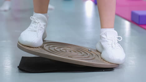 close-up leg view of person in white sneakers stepping on balance board, focusing on stability, blurred background shows another person legs and gym equipment on the ground