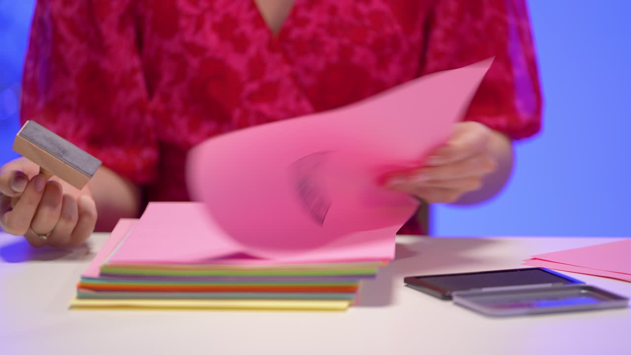 A close-up shows a woman in a red dress using a wooden stamp on pink paper sheets, surrounded by colorful documents and office tools, all within a vibrant and creative workspace setup