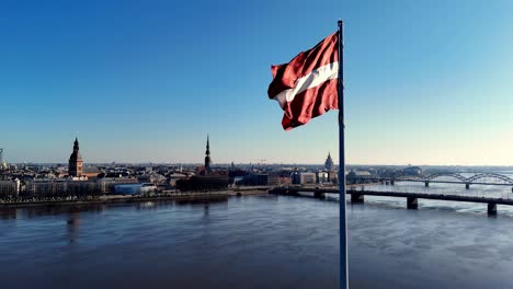 a red and white latvian flag is flying over a riga oldtown city with a river in the background