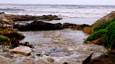 murky rainwater runs into rough sea on coastline from drainage system, telephoto