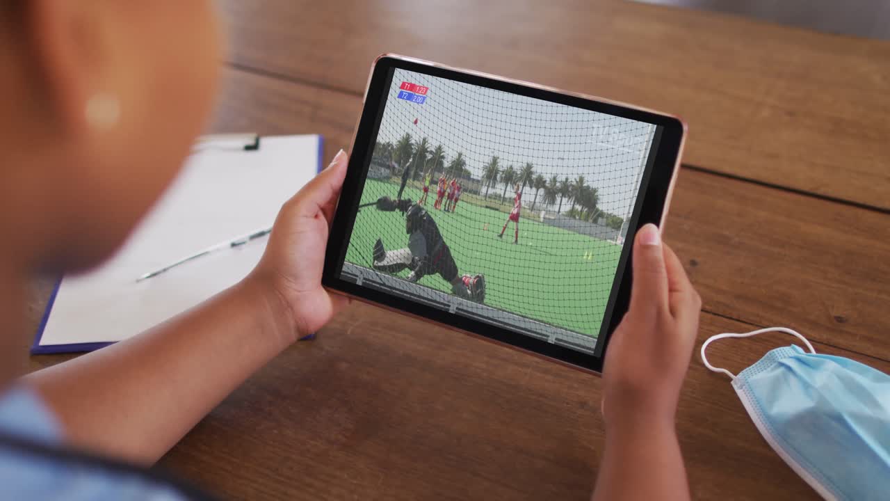 태블릿에서 하키 경기를 시청하는 얼굴 마스크와 함께 테이블에 앉아있는 여성의 복합