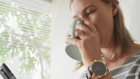 mujer caucásica bebiendo café y sonriendo mientras habla por videollamada en un teléfono inteligente en casa