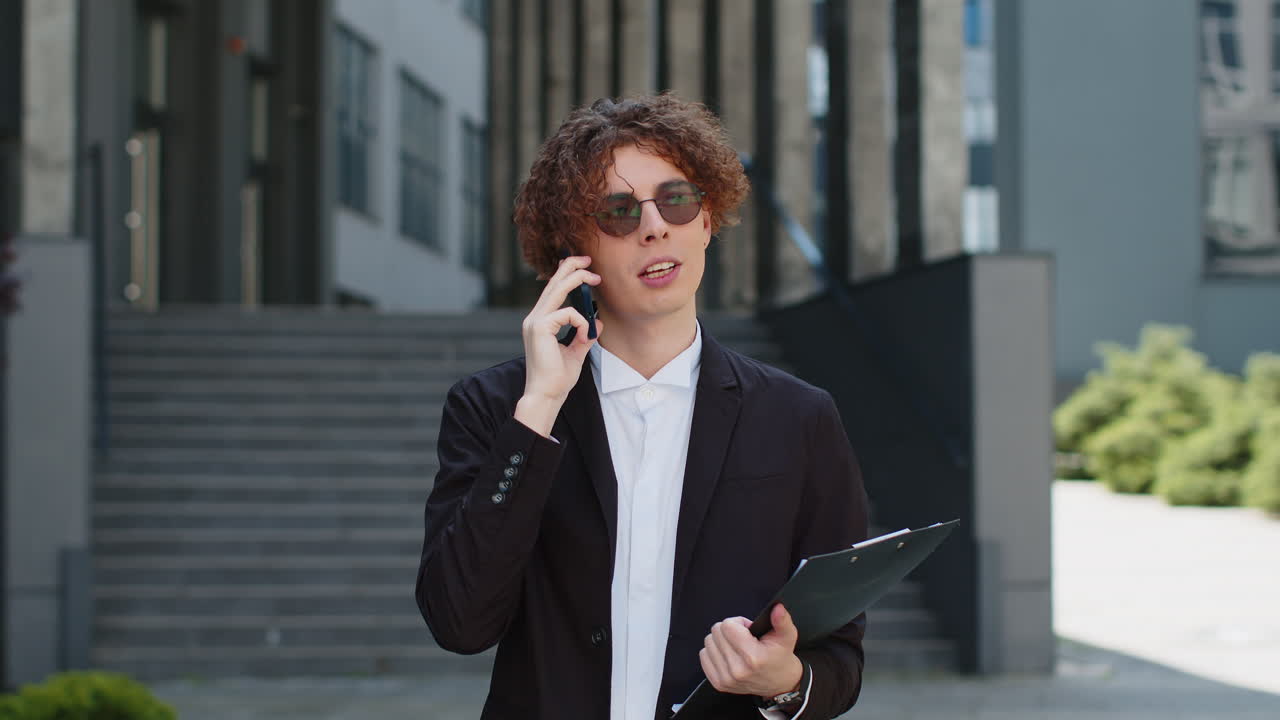 Happy young businessman talking on smartphone on city street near office holding documents clipboard