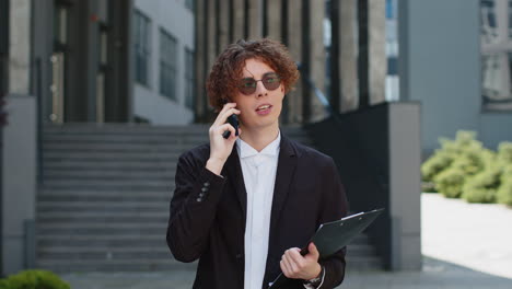 happy young businessman talking on smartphone on city street near office holding documents clipboard