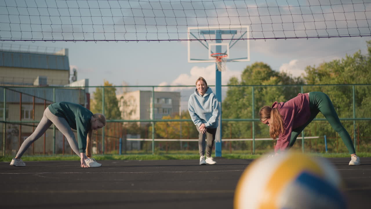 Nærbilde av volleyball på bakken med uskarp bakgrunn som viser idrettsutøvere som tøyer og trener utendørs. En bygning kan sees i det fjerne under en klar himmel